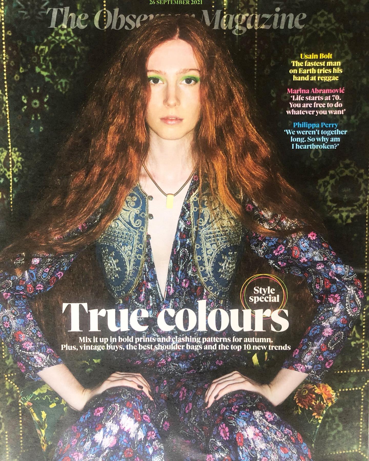 Magazine cover featuring a fashionably dressed adult woman with long red hair in a paisley jumpsuit, posing indoors against a dark backdrop.