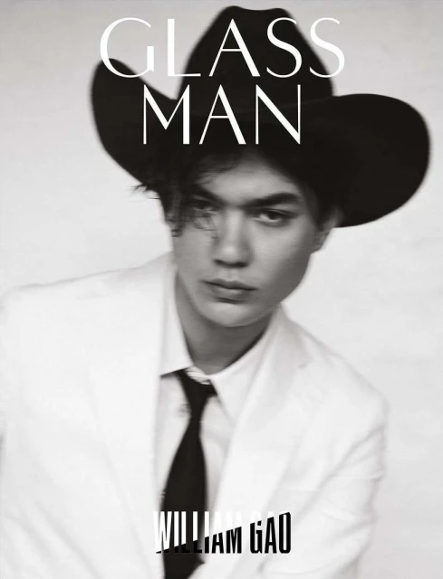 Black and white portrait of a person with a black cowboy hat, white suit jacket, and black tie looking at the camera. 