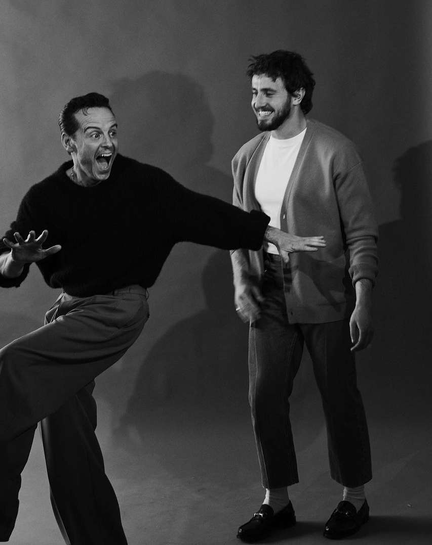 Two adult men pose indoors; one with a wide-eyed expression and a black sweater, the other smiling with a cardigan and a t-shirt.
