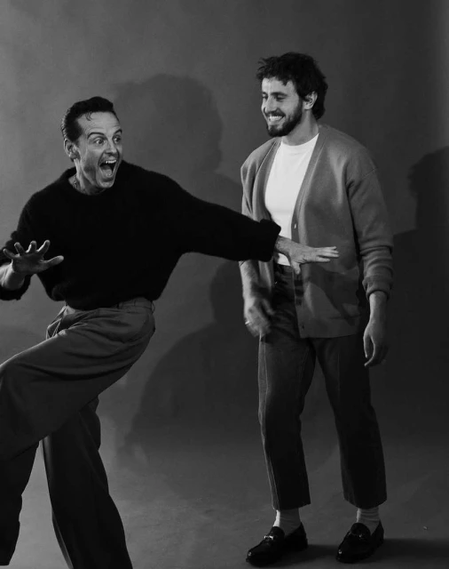 Two adult men pose indoors; one with a wide-eyed expression and a black sweater, the other smiling with a cardigan and a t-shirt.