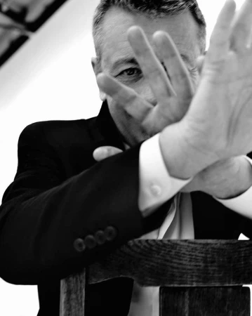 Monochrome portrait of an adult male in a suit, partially obscuring his face with his hand, leaning on a wooden chair.