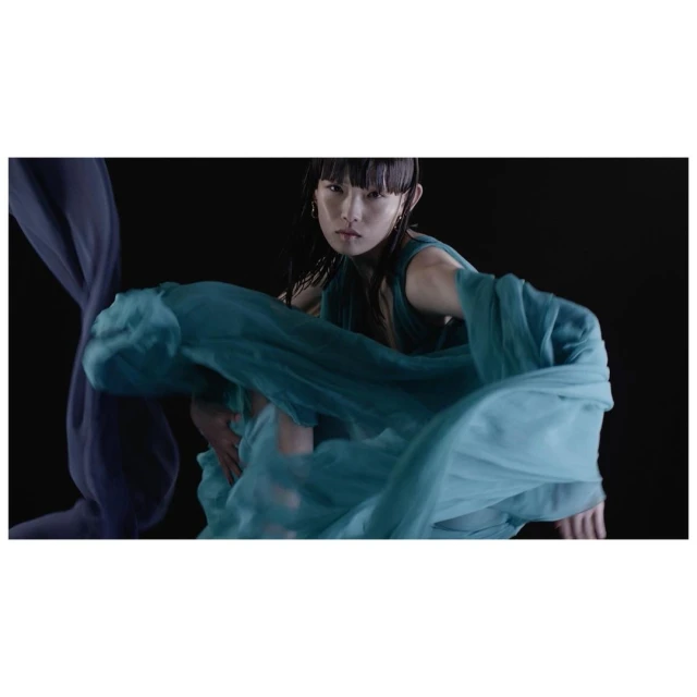 Woman in motion wearing a teal dress and jewelry, posing in front of a black background with translucent blue fabric.