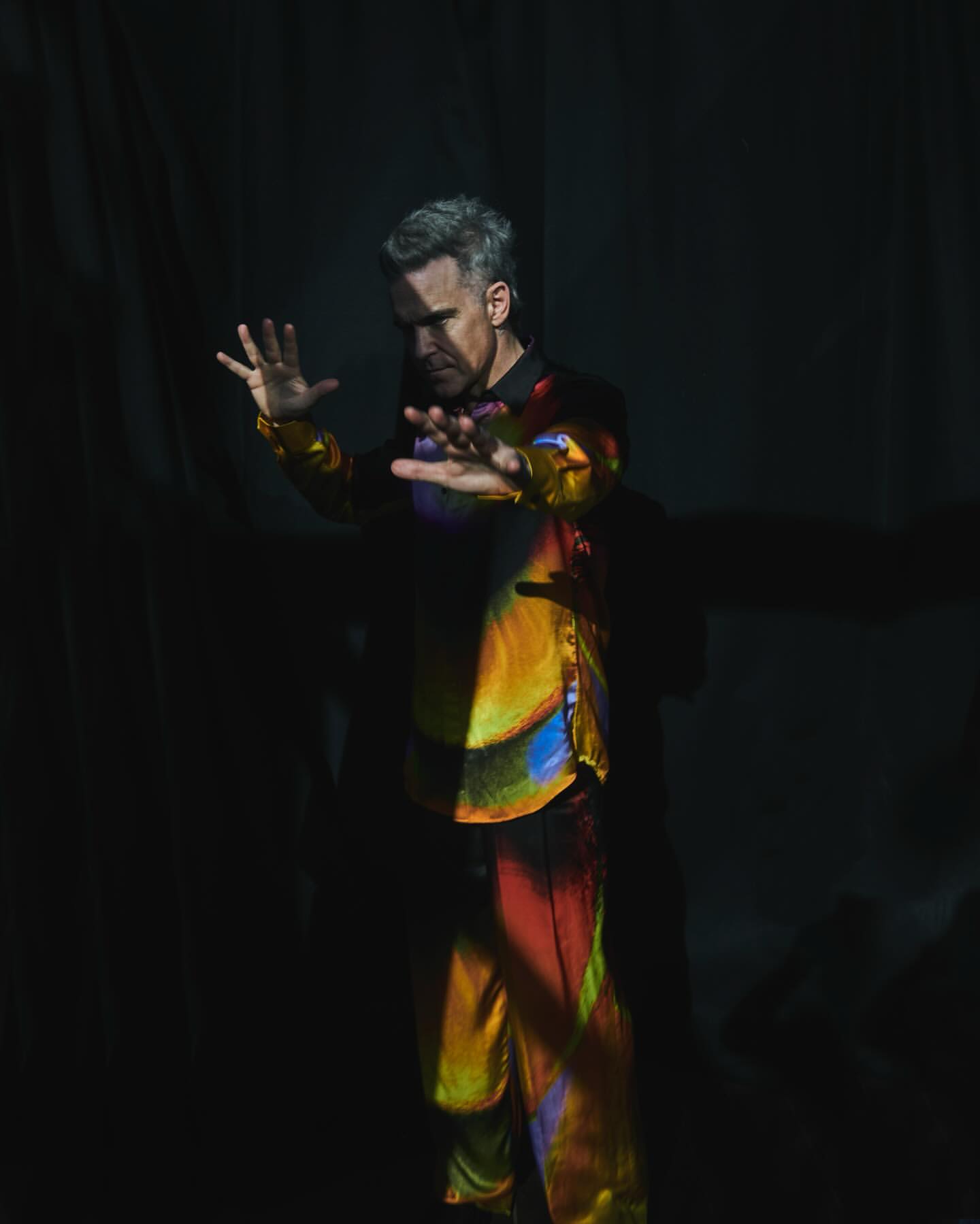 Man in iridescent suit poses in spotlight against dark background. Fashion and creative imagery for advertising and editorial use.
