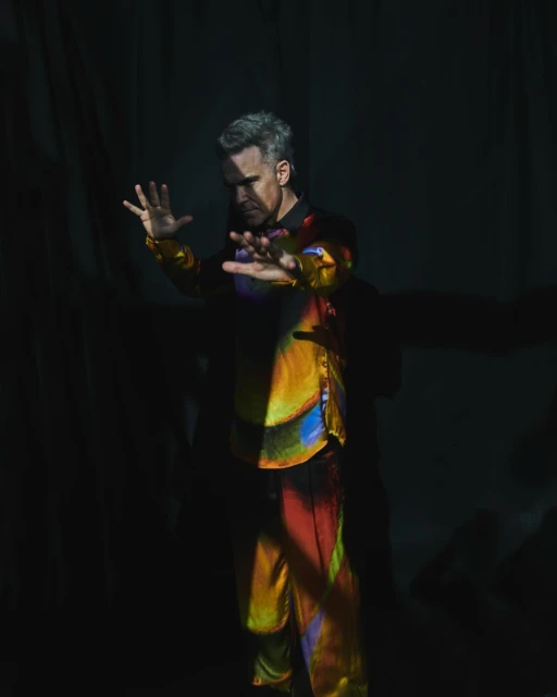 Man in iridescent suit poses in spotlight against dark background. Fashion and creative imagery for advertising and editorial use.