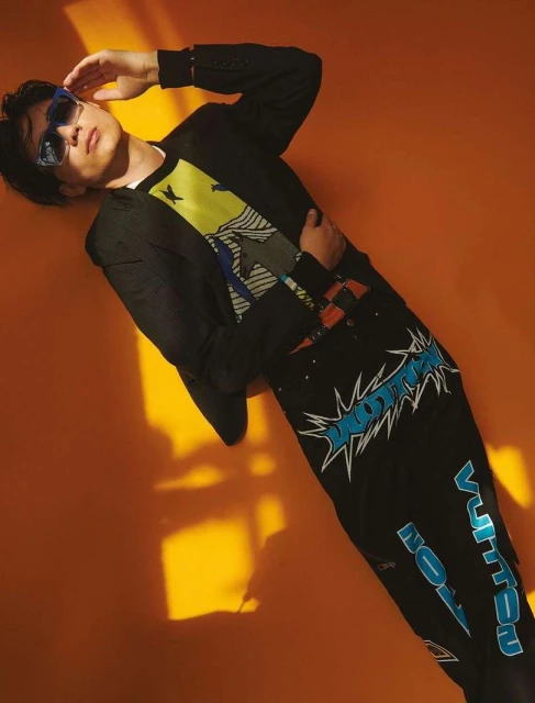 Asian man in black suit and graphic shirt poses against orange background wearing sunglasses and designer pants with logo details.