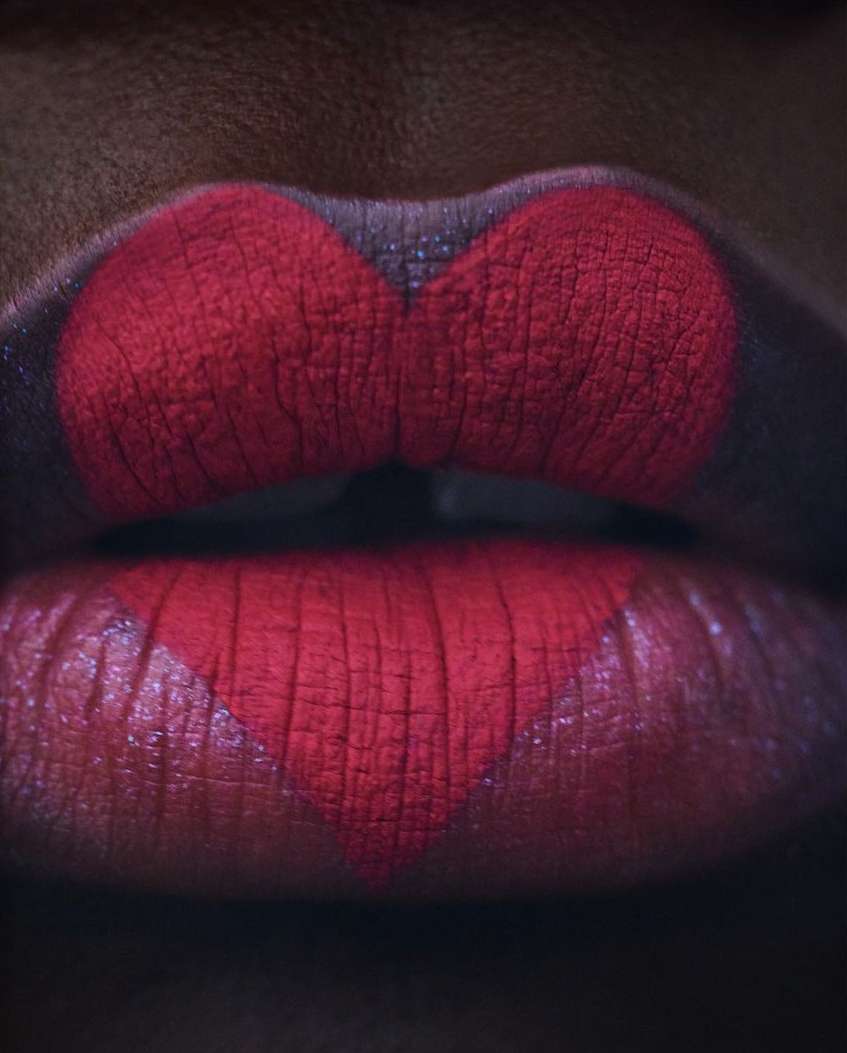 Close-up of a woman's lips with red lipstick forming a heart shape on the lower lip against a dark complexion background.