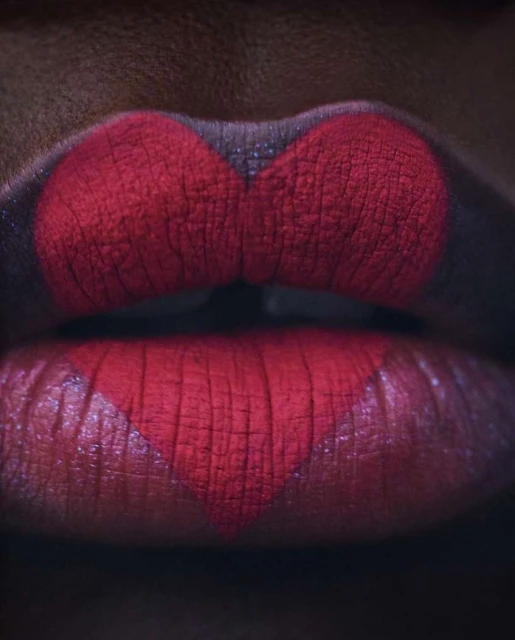 Close-up of a woman's lips with red lipstick forming a heart shape on the lower lip against a dark complexion background.