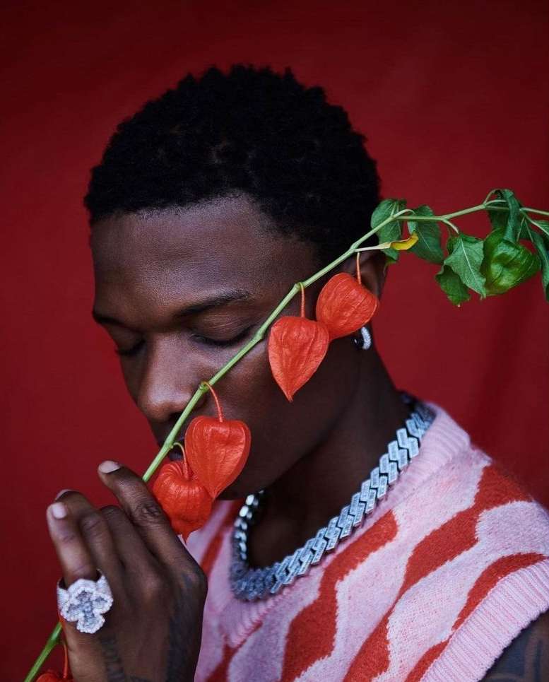 Close-up portrait of an adult male with short black hair, holding orange physalis plant against a solid red backdrop. He wears jewelry and pink top.
