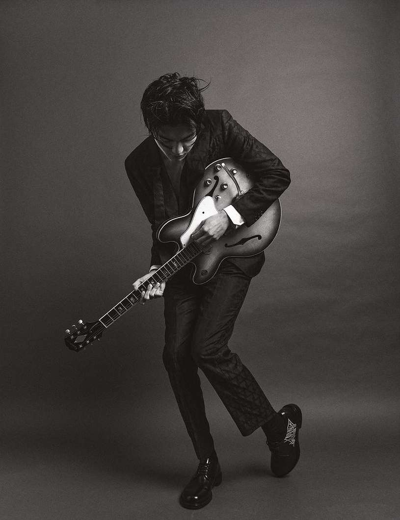 Black and white portrait of an adult male musician playing an electric guitar in a photo studio with dark neutral background.