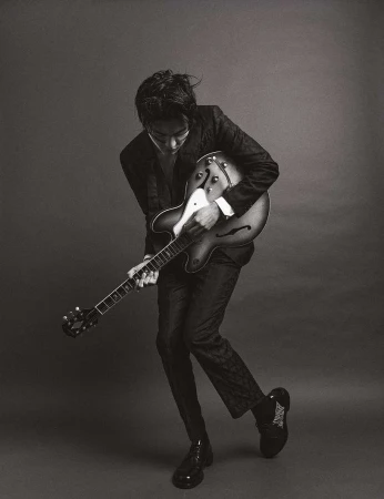 Black and white portrait of an adult male musician playing an electric guitar in a photo studio with dark neutral background.