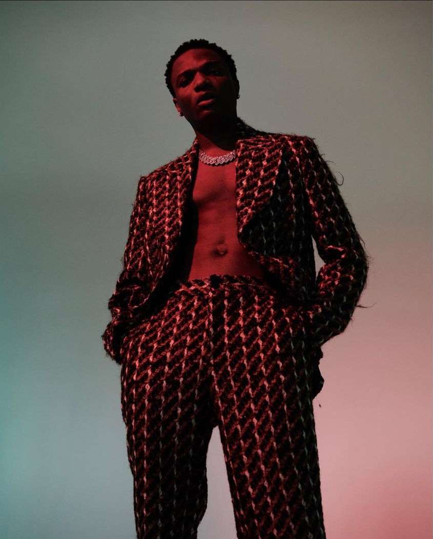 Low-angle studio shot of a man with bare chest wearing a patterned suit, looking at the camera, with a gradient teal and red background.