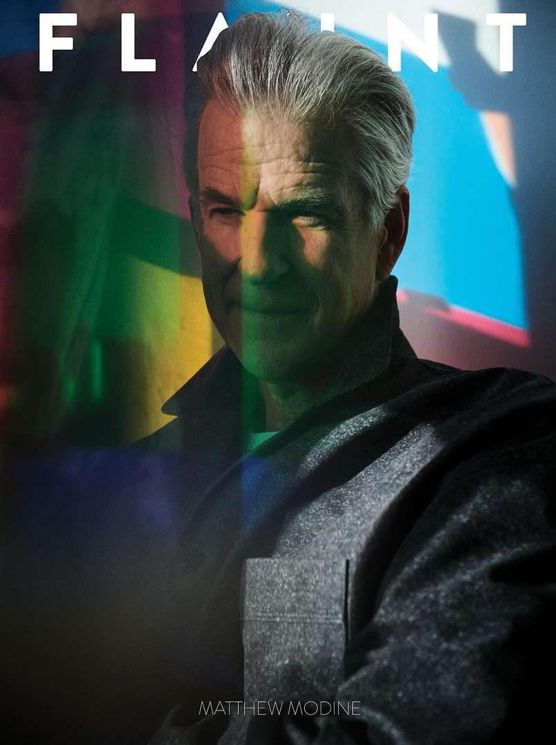 Portrait of adult man with gray hair wearing a gray coat in front of a colorful background with the word 'FLAUNT' and 'MATTHEW MODINE'.