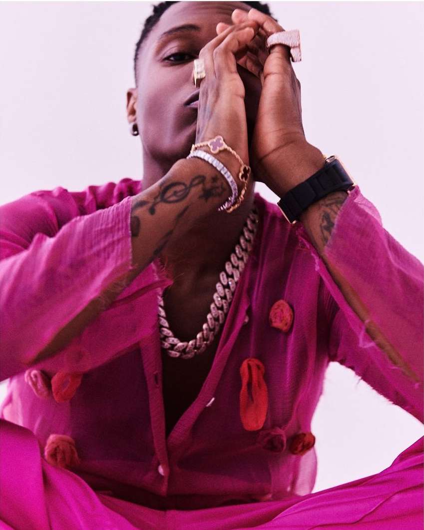 Adult male in magenta outfit poses in a studio, adorned with bracelets, rings, and a chain. His hands are raised in a prayer pose.