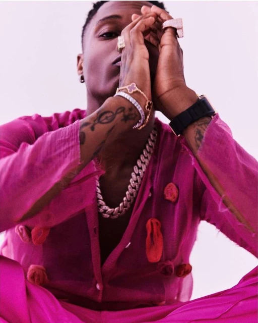 Adult male in magenta outfit poses in a studio, adorned with bracelets, rings, and a chain. His hands are raised in a prayer pose.