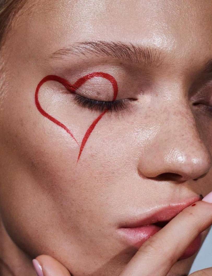 Close-up portrait of a woman's face with a painted red heart around her eye, finger to lips, for beauty, fashion, or editorial use.