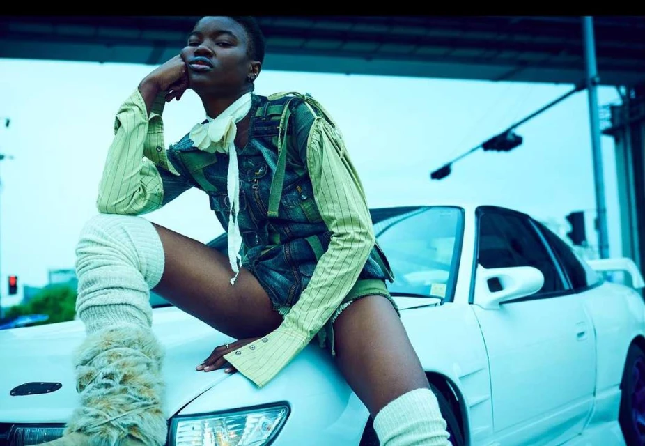 Stylish woman in vintage-style clothing poses on a white car in an urban setting with overpass background in soft blue-toned daylight.