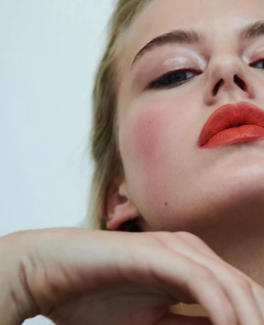 Close-up of a young fair-skinned woman with blond hair and coral lipstick, posing against a white background in a beauty portrait.