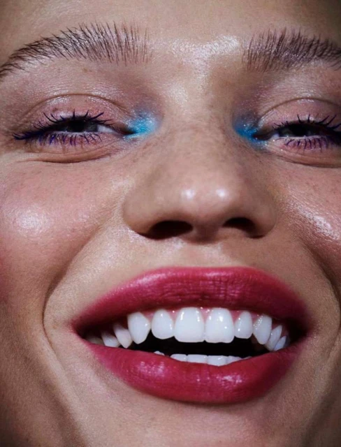 Close-up of a woman's smiling face showcasing vibrant makeup with red lipstick and bright blue eyeshadow, highlighting a beauty aesthetic.