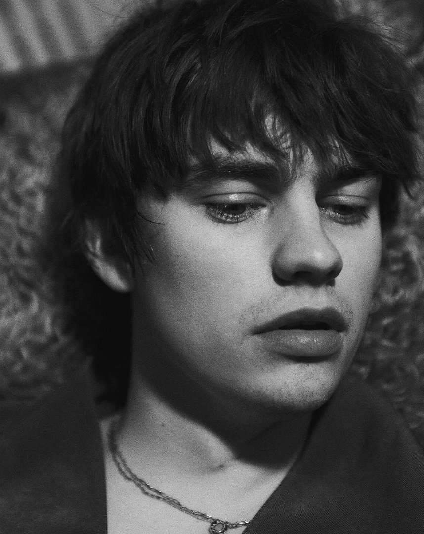 Close-up black and white portrait of a young man with dark tousled hair and a thin chain necklace by Jason Hetherington