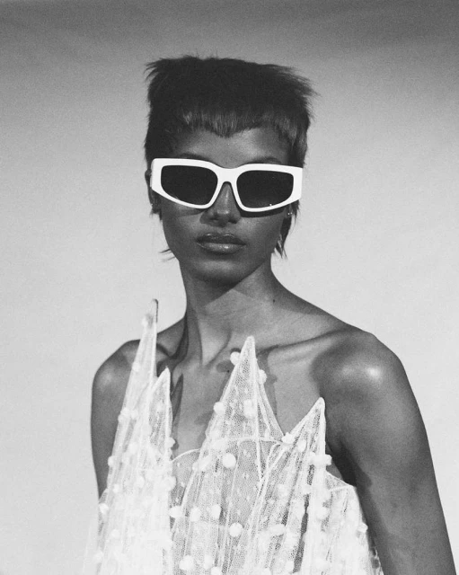 Close-up shot of a woman wearing oversized white sunglasses and a decorative translucent white dress with raised geometric shapes.