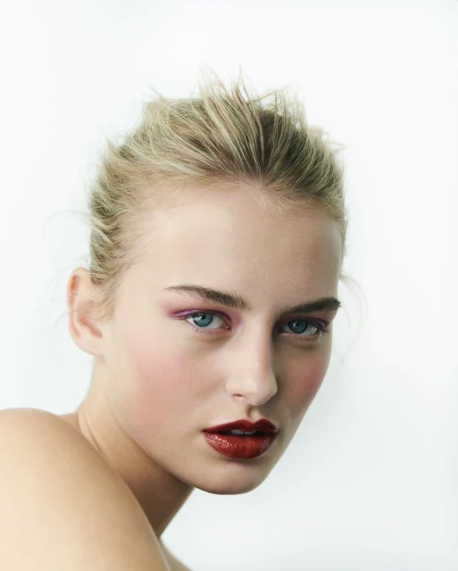 Close-up portrait of a fair-skinned woman with blonde hair pulled back, wearing pink eyeshadow and dark red lipstick against a white background.