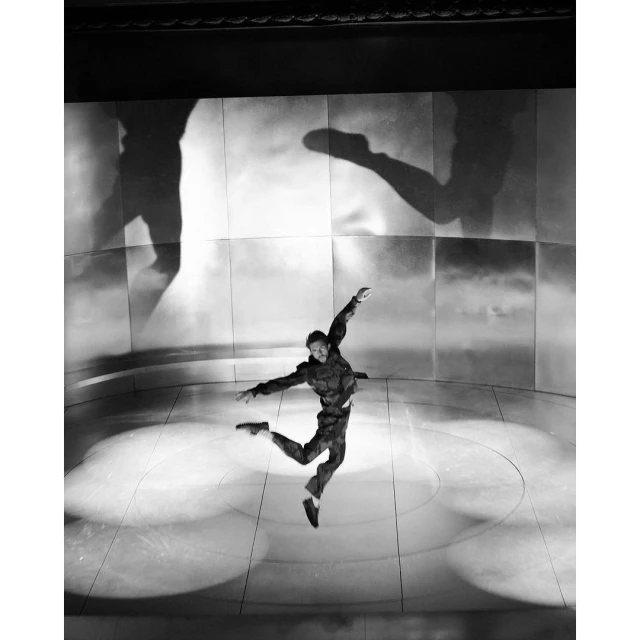 Black and white dance performance photo: A male dancer leaps on stage in front of a backdrop with large shadow projections.
