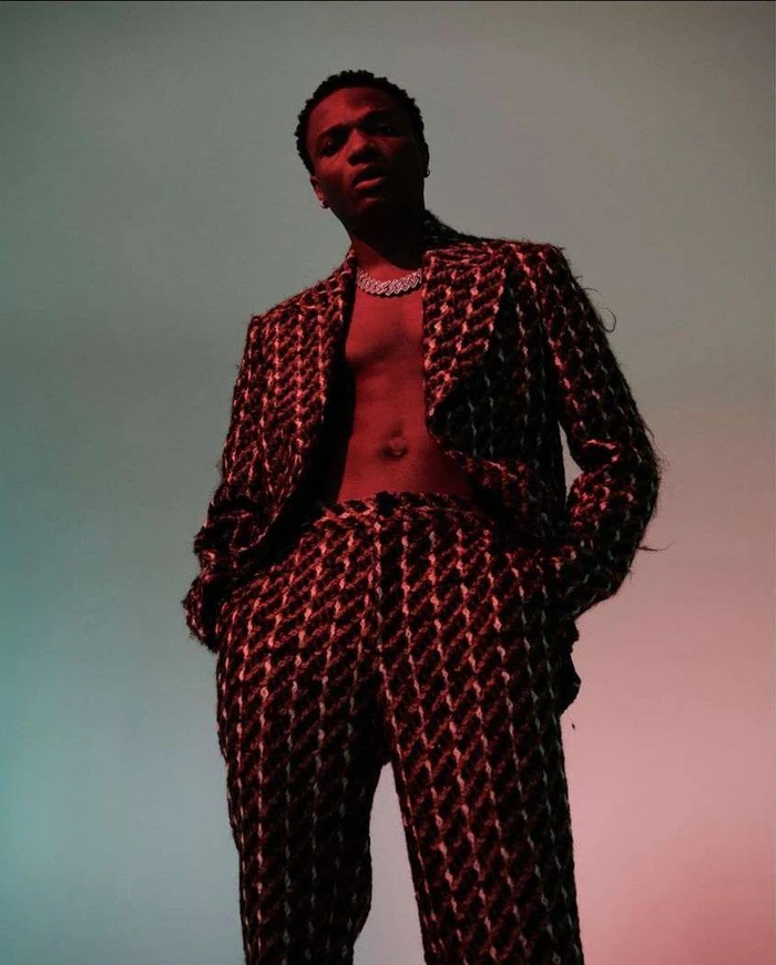 Black man wearing a patterned tweed suit with no shirt underneath, photographed from a low angle against a gradient background.