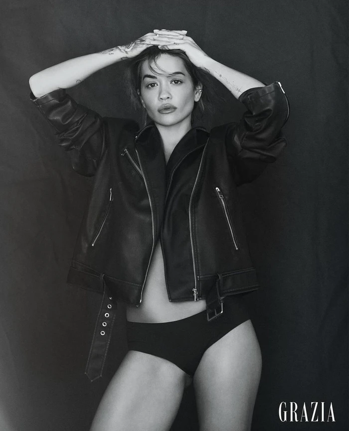 Black and white editorial portrait of Rita Ora wearing an oversized leather jacket and underwear against a dark textured backdrop.