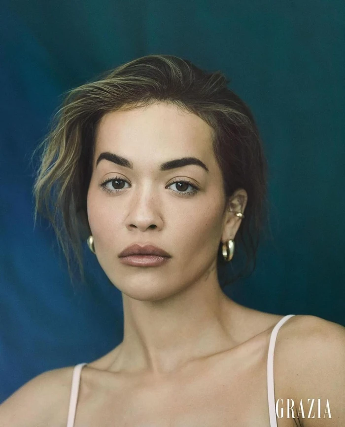 Close-up portrait of Rita Ora with dark hair and neutral makeup, wearing gold hoop earrings against a teal background