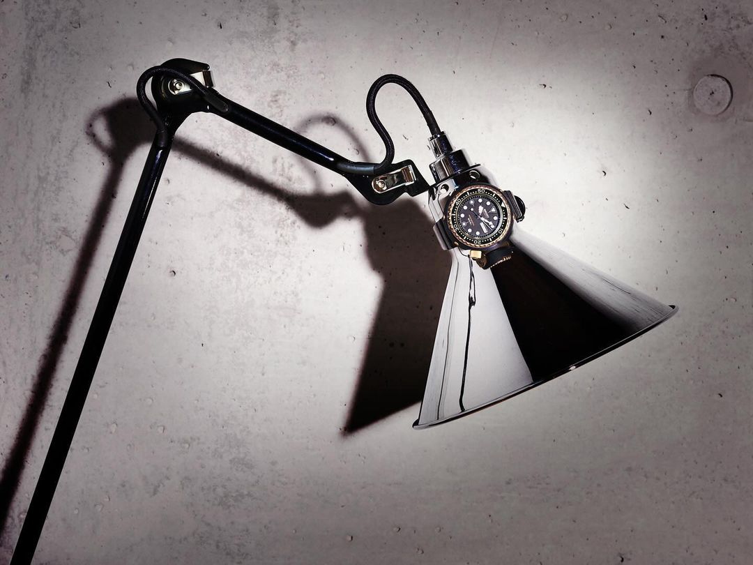 Dramatic studio shot of a black metal articulated desk lamp illuminating a black-faced wristwatch against a concrete wall backdrop.