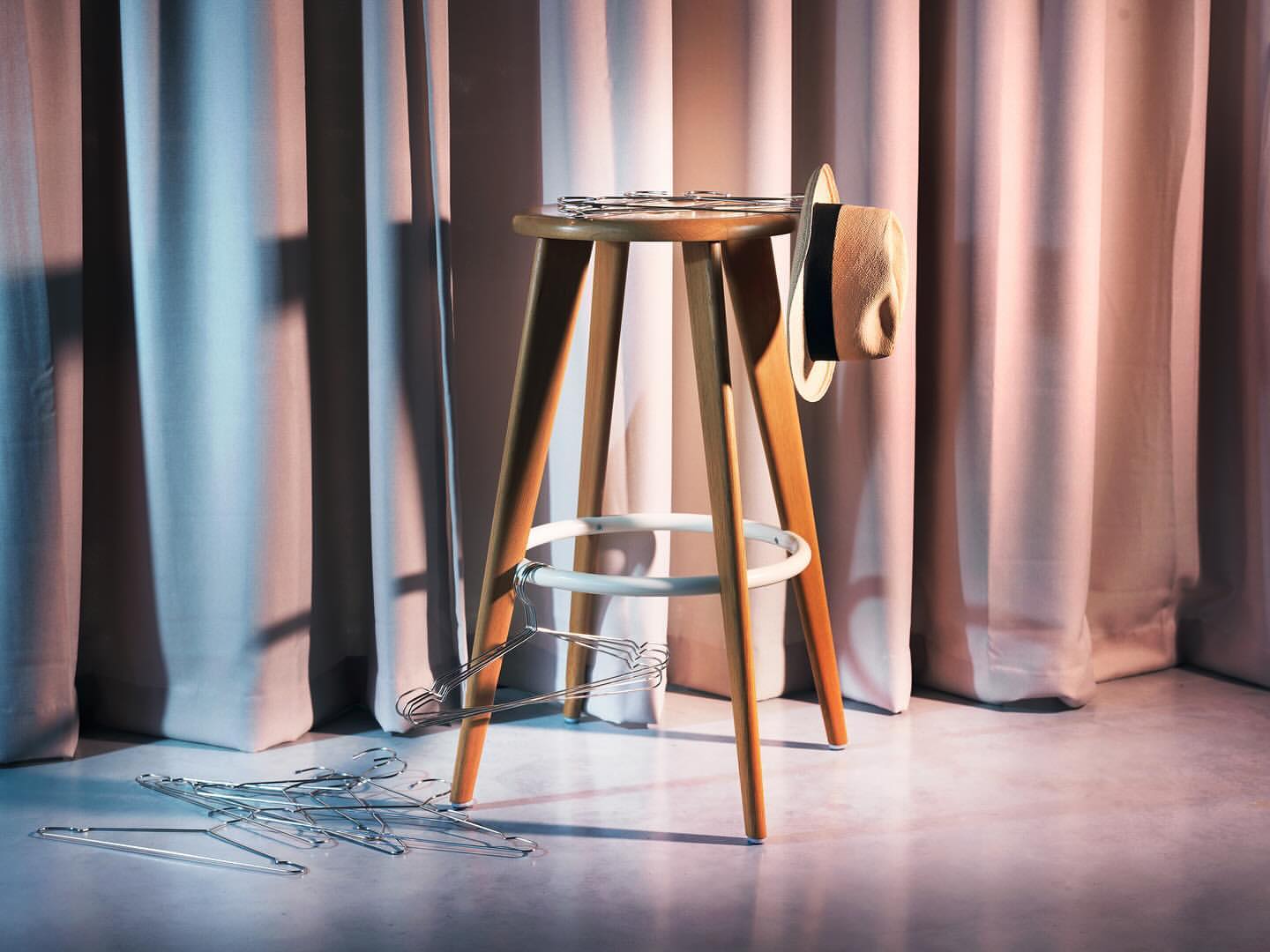 A wooden stool with a hat and clothes hangers stands against draped curtains, illuminated with a mix of warm and cool lighting.