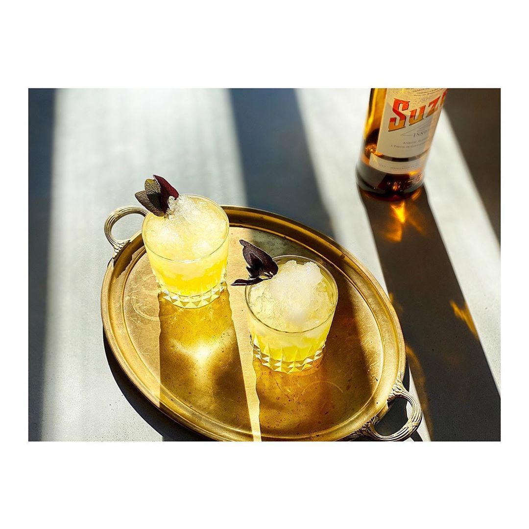 Two yellow cocktails with crushed ice and basil garnish on a gold tray with a bottle of liquor and sunlight in the background.