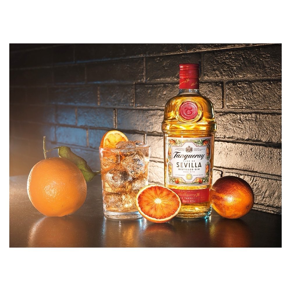 Tanqueray Sevilla gin bottle next to a glass of gin and tonic with orange slices and blood oranges against a dark background.