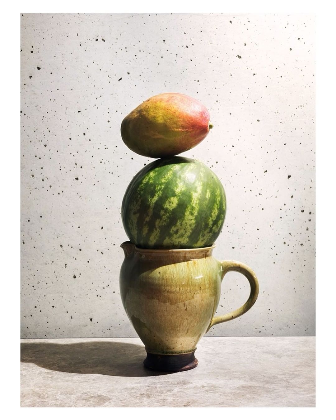 Still life featuring a mango atop a watermelon, which rests on a rustic ceramic pitcher against a textured gray backdrop.