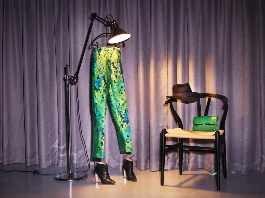 Fashion still life featuring green floral pants hanging from a floor lamp, styled with ankle boots, purse, and hat against gray curtains.