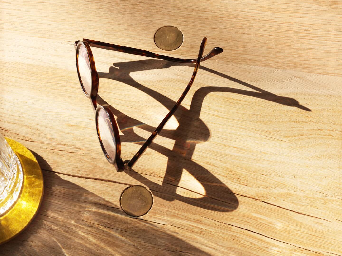 Tortoiseshell glasses resting on a wooden surface with a round, gold-colored object, captured in warm, diffused sunlight casting long shadows.
