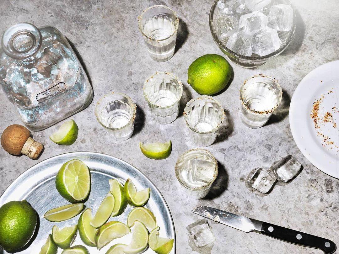 Overhead shot of tequila bottle, shot glasses with salt rims, ice, limes, and a knife, set on a grey surface, food and beverage.