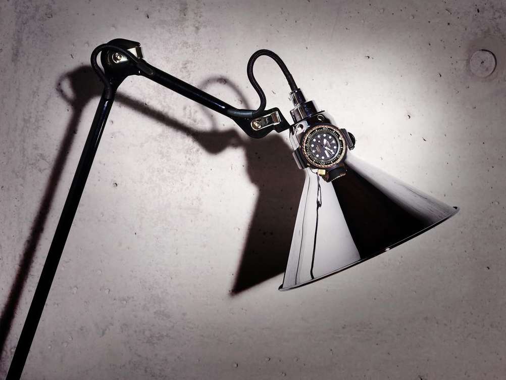 Wristwatch mounted on the shade of an adjustable lamp, illuminating the timepiece against a concrete wall backdrop. Dark and industrial.