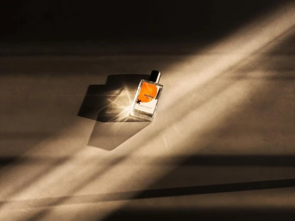 Perfume bottle with orange label on surface with patterned lighting and shadows, product shot, beauty concept, fragrance marketing.