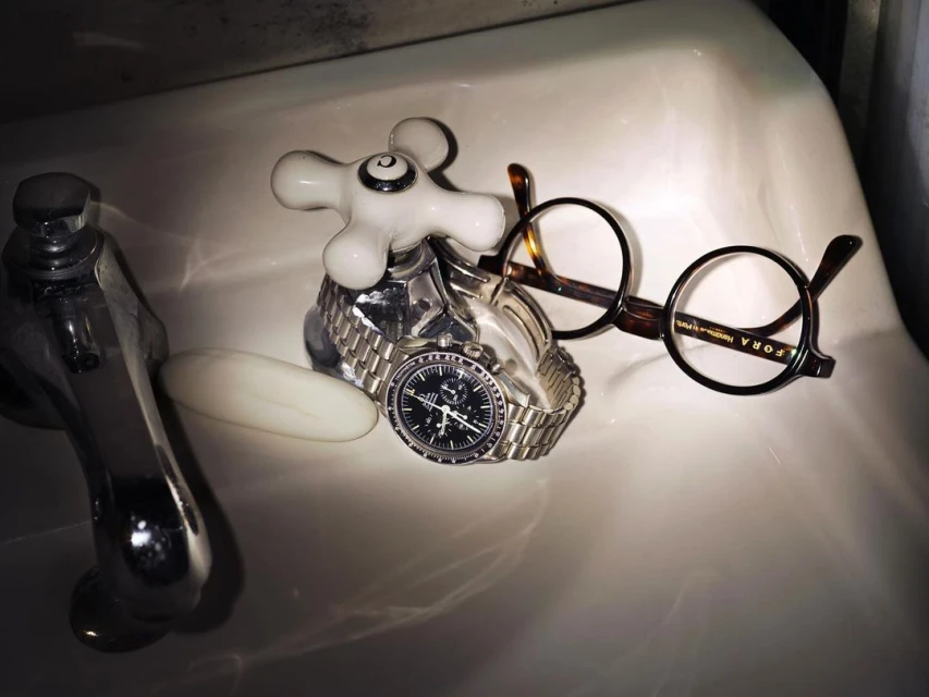 Still life of a silver wristwatch on a faucet, eyeglasses, and a bar of soap, all arranged in a white porcelain sink.