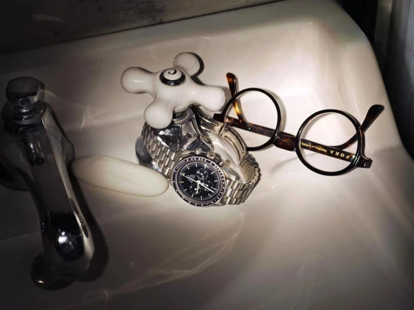 Still life of a silver wristwatch on a faucet, eyeglasses, and a bar of soap, all arranged in a white porcelain sink.