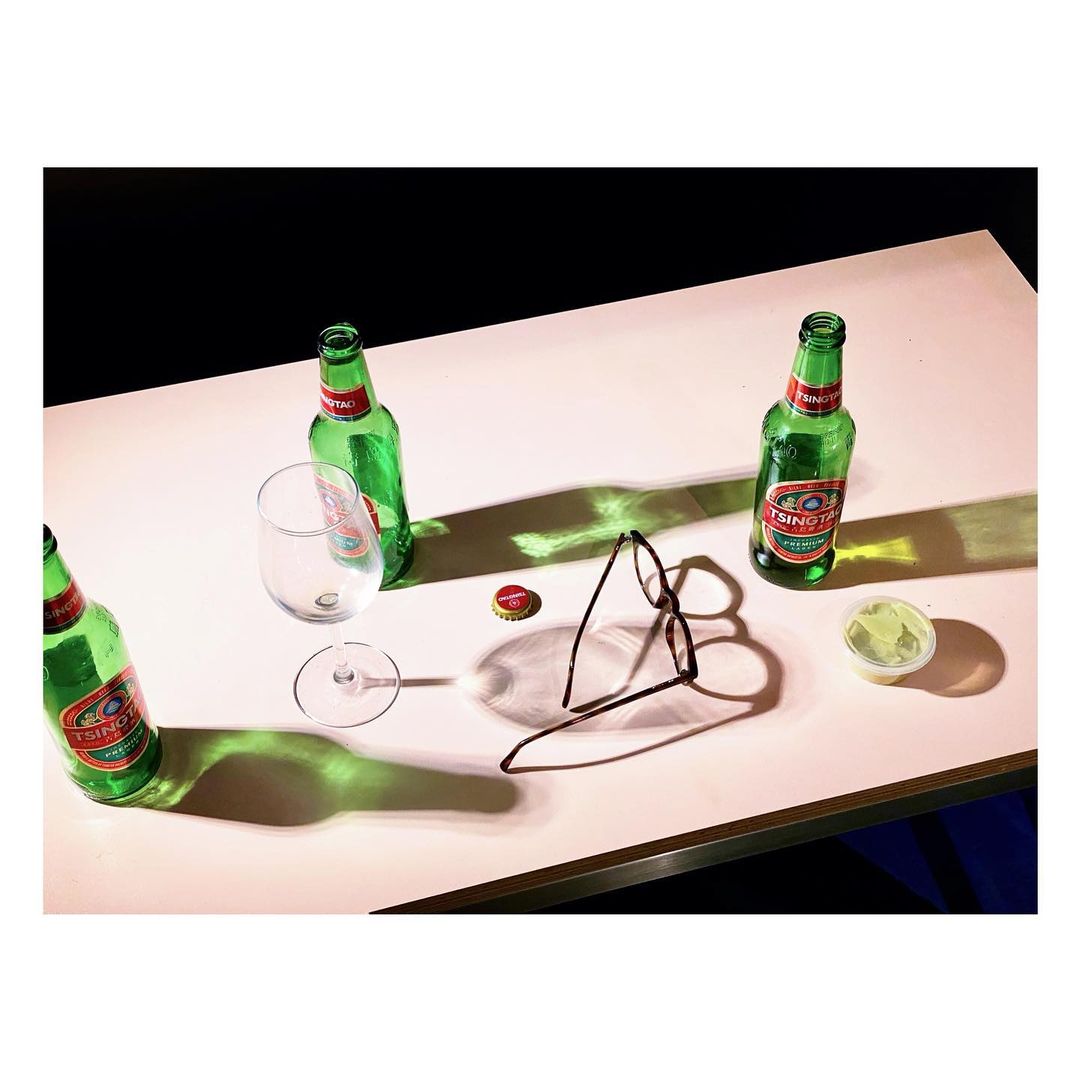 Overhead shot of Tsingtao beer bottles, a stemmed glass, a bottle cap, eyeglasses, and a condiment cup on a pink table against a dark background.