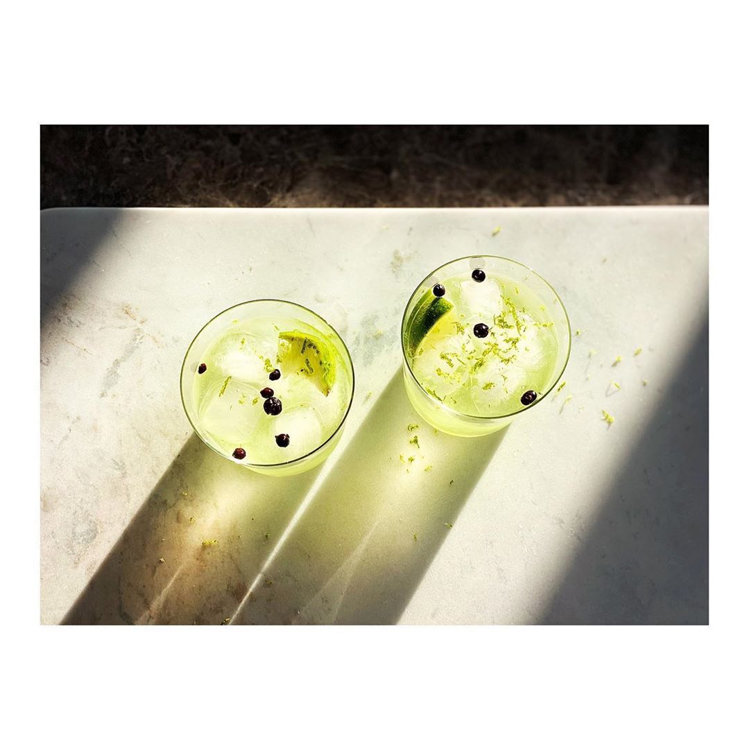 Two glasses of a lime-colored cocktail with ice, lime wedges, and black peppercorns on a marble surface with shadows.