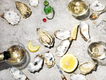 Flat lay photograph of fresh shucked oysters, lemon wedges, glasses of white wine, a small green sauce bottle, and an oyster knife.
