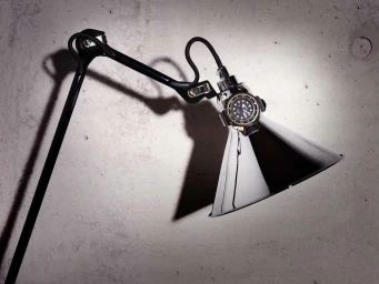 Close-up of a black adjustable lamp with a metallic cone shade and a wristwatch mounted on it against a concrete wall