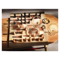 Overhead view of a chess set on a marble board with two coupe glasses and jewellery on a wooden table under direct sunlight.