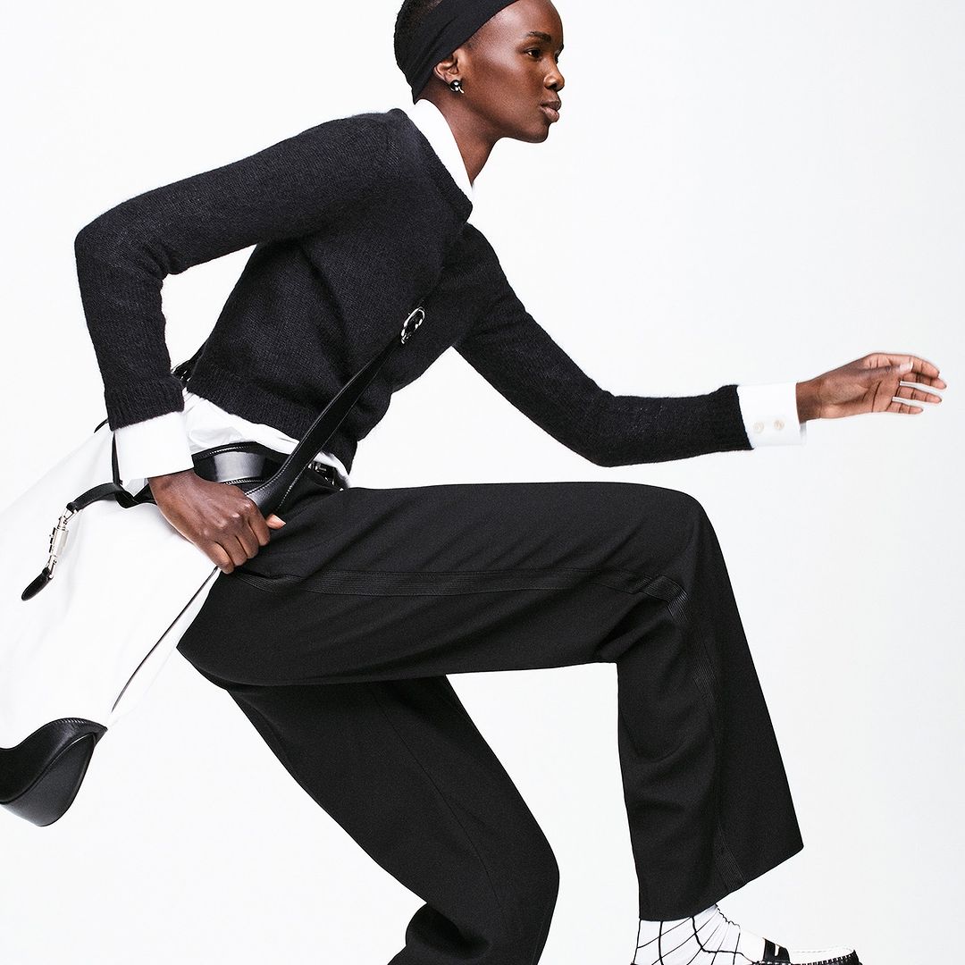 Fashionable black female model in motion, wearing a dark sweater, wide-leg trousers, and sandals against a plain white backdrop in a studio setting.