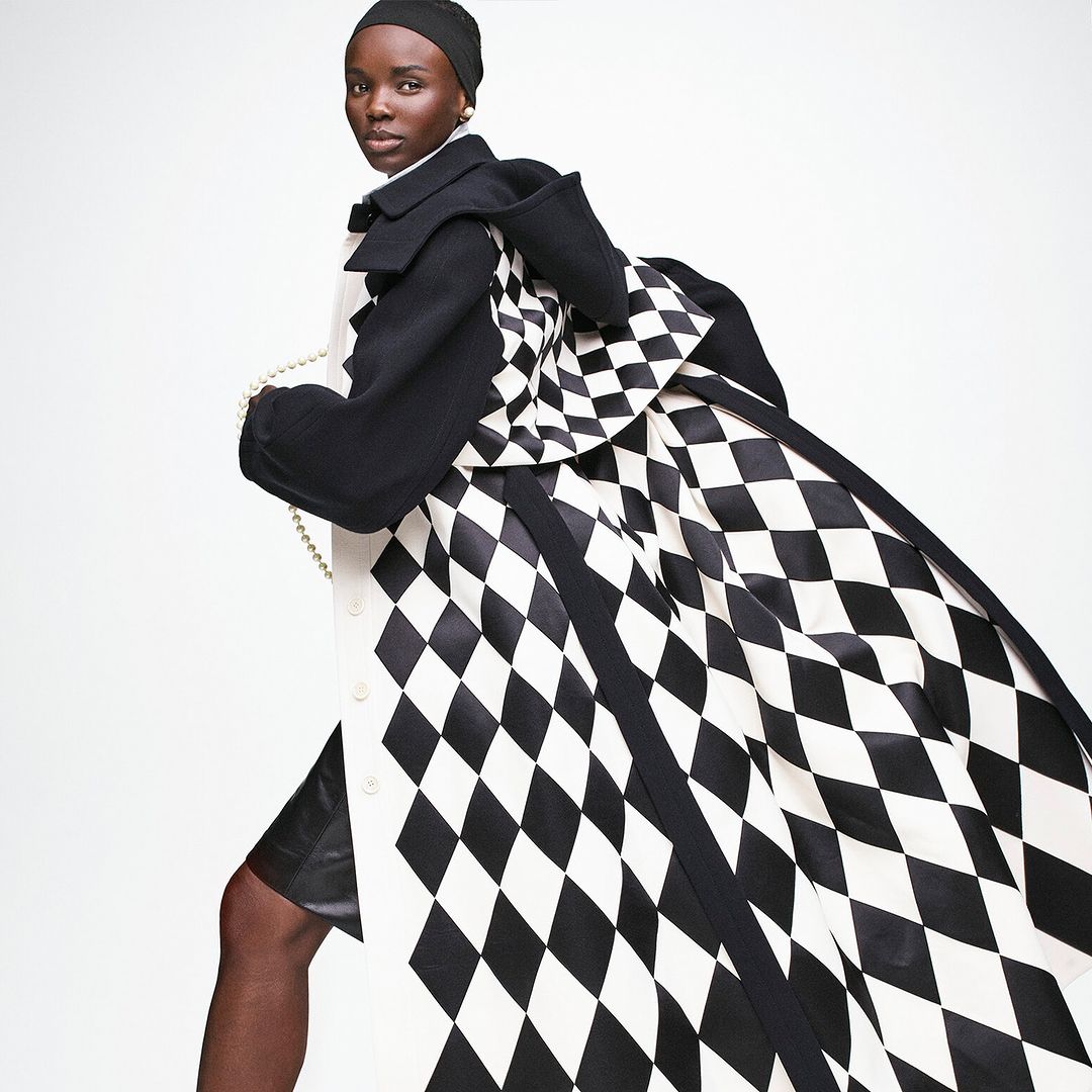 Fashion-forward, dark-skinned woman strikes a pose in a black and white patterned coat, black headscarf, and accessories against a white backdrop.