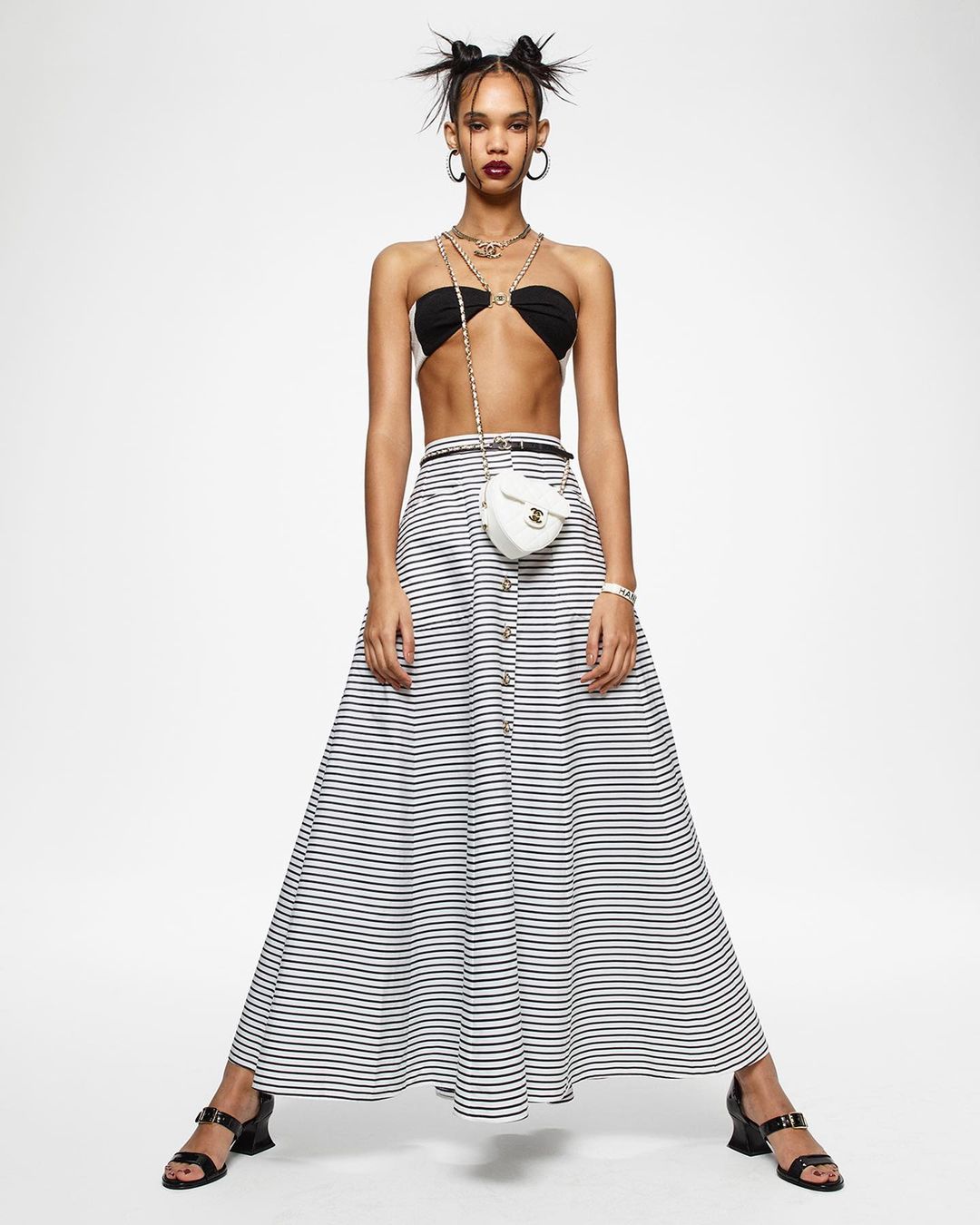 Fashion model posing in a strapless top, striped skirt, and strappy sandals against a white backdrop in a studio environment.