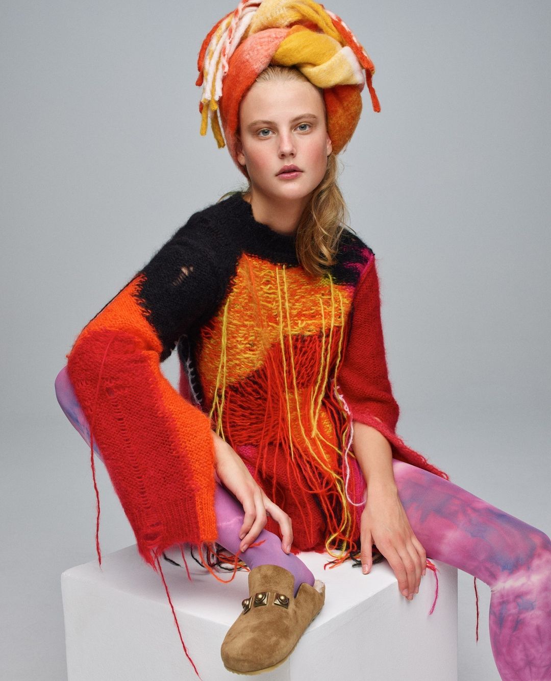 Fashionable young adult woman in colorful knit sweater and head wrap seated on white cube against grey backdrop in studio shot.